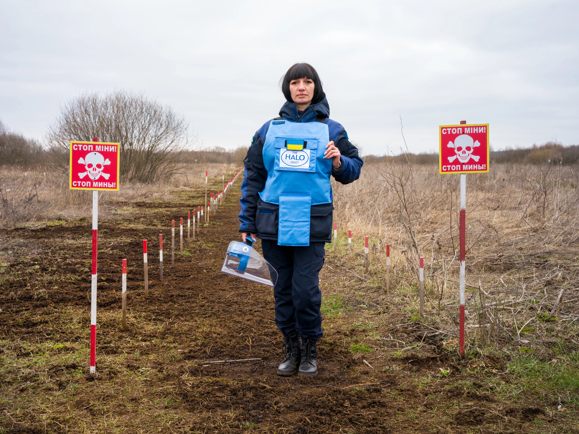 Жінка стоїть на сухій траві у полі між двома рядами червоно-білих стовпів, що тягнуться позаду неї, на задньому плані видніються кущі та дерева, небо тьмяне і хмарне. Попереду кожної лінії стовпів є червоний знак мінної небезпеки з білою позначкою у вигляді черепа та схрещених кісток. Жінка одягнена в чорні черевики, темно-сині водонепроникні штани та куртку в тон, світло-блакитний захисний жилет із логотипом Halo Trust у центрі. У неї темне волосся з чубчиком, що прикриває чоло, в правій руці вона тримає прозору захисну маску. На довгих нігтях у неї такий же синій акриловий лак. Це Олександра Євдокимова, керівниця групи з розмінування організації HALO Trust.