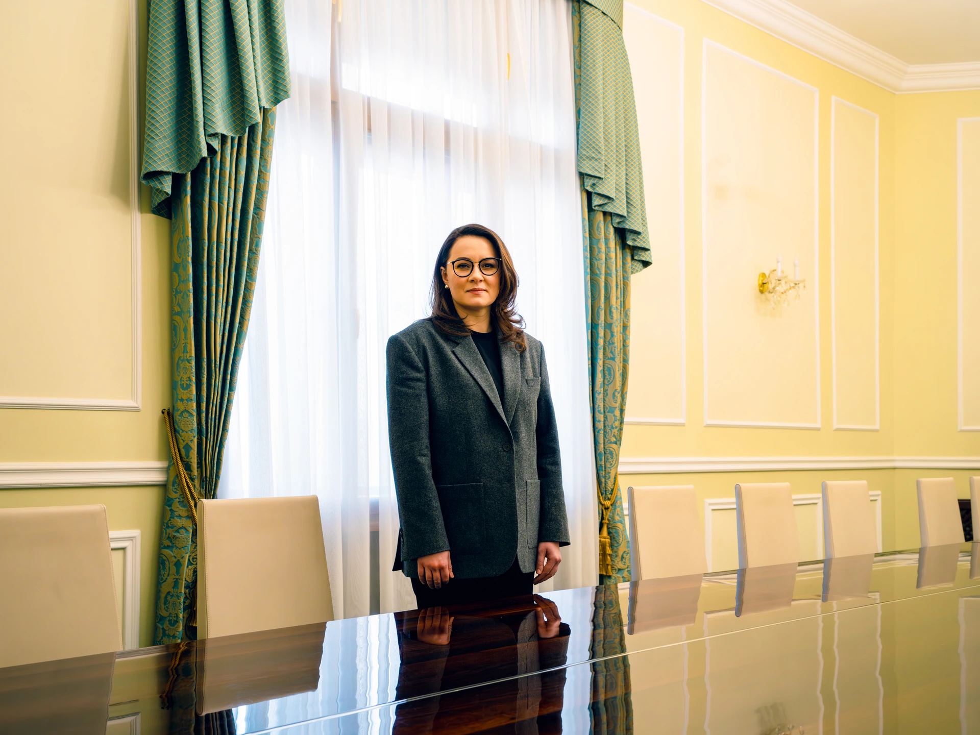 Жінка стоїть у добре освітленій кімнаті з жовтими стінами, та обрамленими у білий колір фальш-панелями та ліпними прикрасами. За нею — вікно з розсунутими зеленими гардинами з візерунчастими золотими прикрасами, а перед нею — великий і блискучий лакований стіл, за яким видніються стільниці кількох бежевих стільців. У неї пряме каштанове волосся до плечей, вона носить сережки, окуляри в чорній оправі, сірий піджак на ґудзиках, її руки опущені вздовж тіла. Це Юлія Свириденко, міністр економіки України, а також перший віцепрем'єр-міністр України.